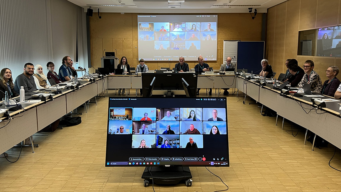 Meeting with participants seated around a conference table and remote attendees joining via video screens.